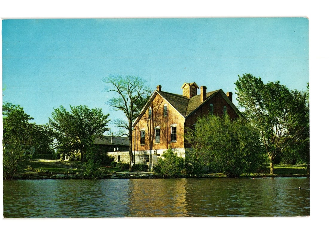 Nauvoo House, Historic Brick Building by Water in Nauvoo, Illinois, a Vintage Postcard: A color photographic postcard depicting Nauvoo House in Nauvoo, Illinois. The prominent brick building, with a lighter foundation, stands on the far bank of a body of water. It features a gabled roof 