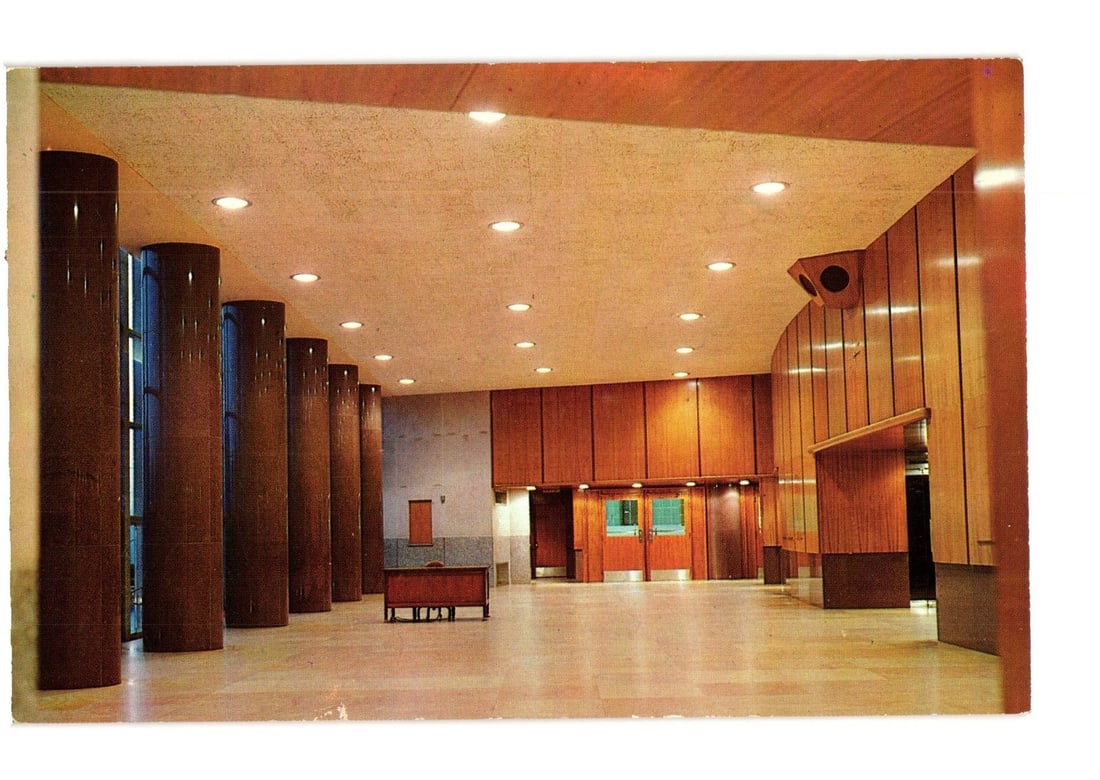 Main Foyer of The Auditorium, Independence, Missouri, mid-20th century architectural postcard: An interior view of a large, modern foyer or lobby, characterized by its distinctive architectural design. The space features a series of dark, cylindrical columns along the left side, paralleling a l