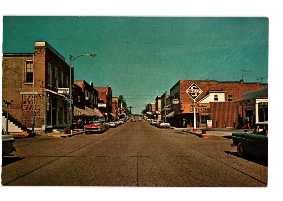 Main Street, Troy, Missouri Small Town View with Vintage Cars and Brick Buildings Color Postcard: A vibrant color photograph depicts a main street scene in a small town, viewed from the street level looking into the distance. Brick buildings line both sides of the thoroughfare, with several mid-20