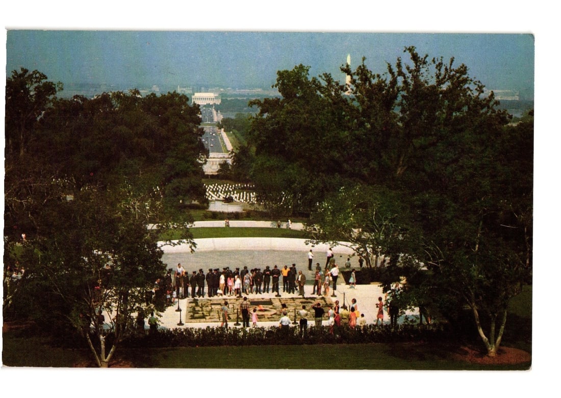 Historic Elevated View of John F. Kennedy Grave, Arlington National Cemetery, Virginia postcard: An elevated view of the John F. Kennedy grave site at Arlington National Cemetery. A group of people is gathered around the central memorial, which features a low wall. Several individuals among the c
