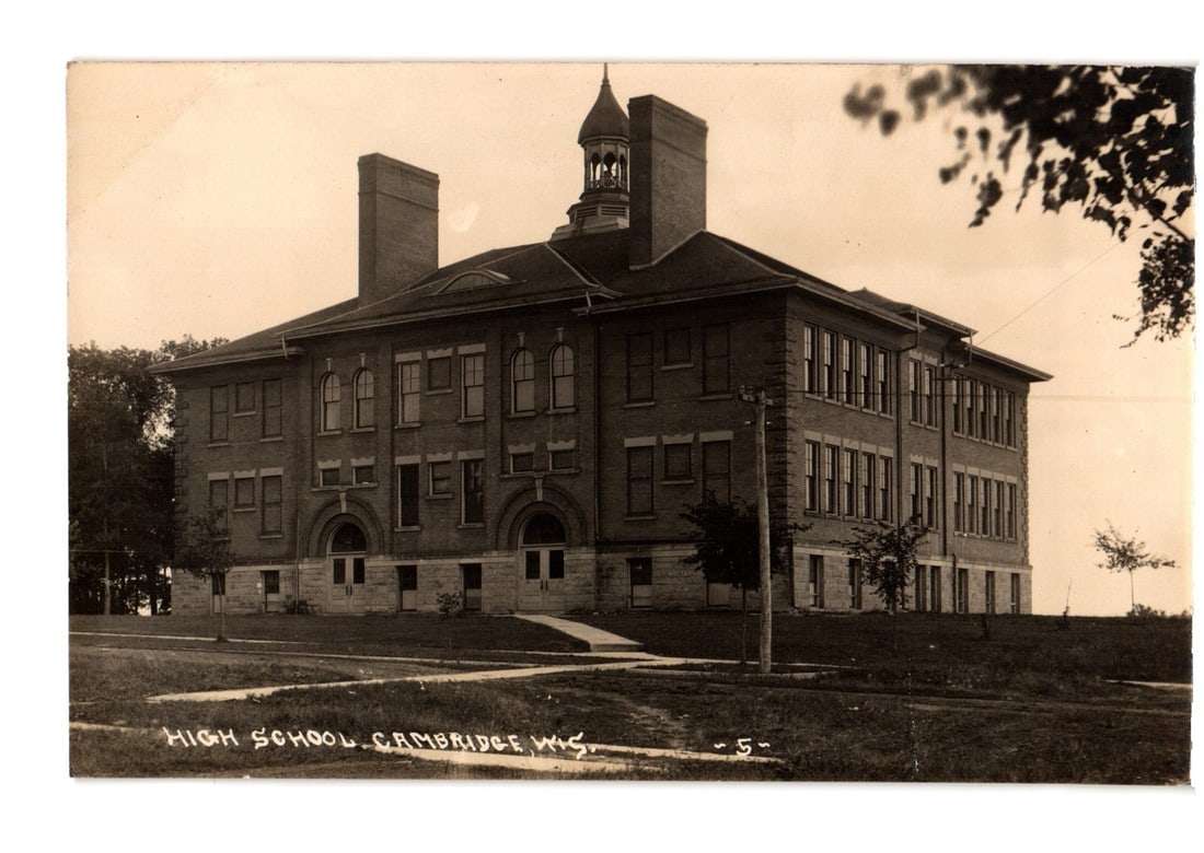 Early 20th century sepia photographic postcard of the High School in Cambridge, Wisconsin.: A three-story brick high school building is depicted from a slightly elevated, angled perspective. The architecture features a central cupola with a bell tower, multiple chimneys, numerous rectangular