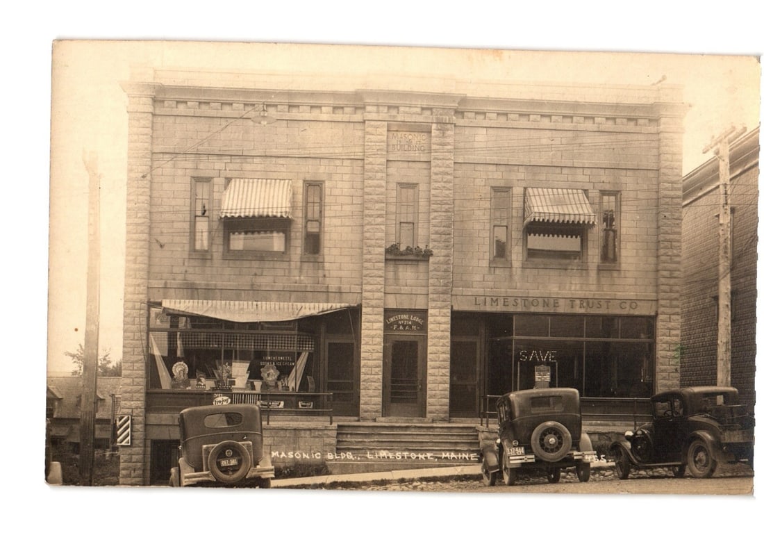 Vintage Postcard: Masonic Building, Limestone Trust Co., with Period Automobiles, Limestone, Maine: A large, two-story stone building features two commercial storefronts on the ground floor and multiple windows above. The facade is constructed of dressed stone blocks, with striped awnings extended o