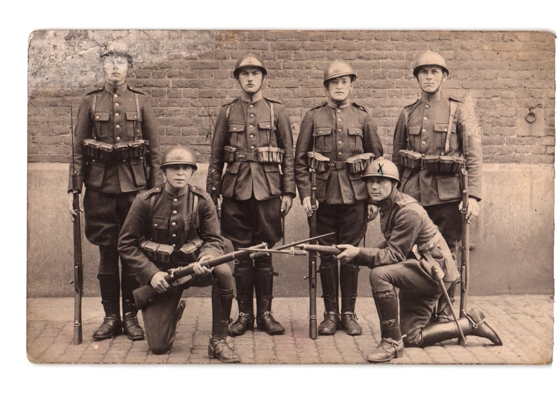 WWI-Era Military Postcard: Six Soldiers in Uniform with Rifles Posing Against Brick Wall.: Six soldiers are depicted in full military uniform, posed against a brick wall background. Three soldiers stand in the rear, while two kneel and one crouches in the foreground. All wear helmets, boots