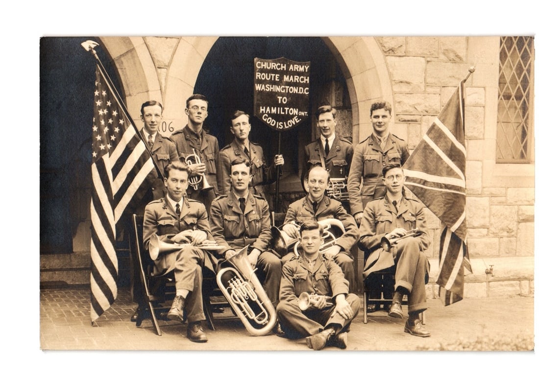 Church Army Route March Band, Washington D.C. to Hamilton, Ontario, Group Portrait postcard: A group portrait photograph depicts ten men in military-style uniforms, positioned outdoors in front of a stone building with an arched entrance. Several men hold brass musical instruments including a