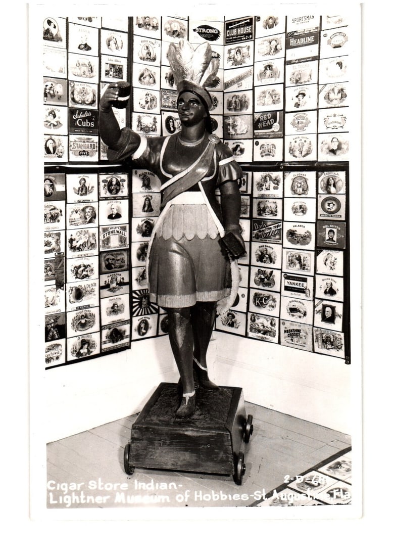 Cigar Store Indian Statue, Lightner Museum of Hobbies, St. Augustine, Florida postcard: A full-length photographic depiction of a carved wooden figure known as a Cigar Store Indian. The female figure, with dark skin, wears a feathered headdress, a tunic, and a sash, holding an object alo
