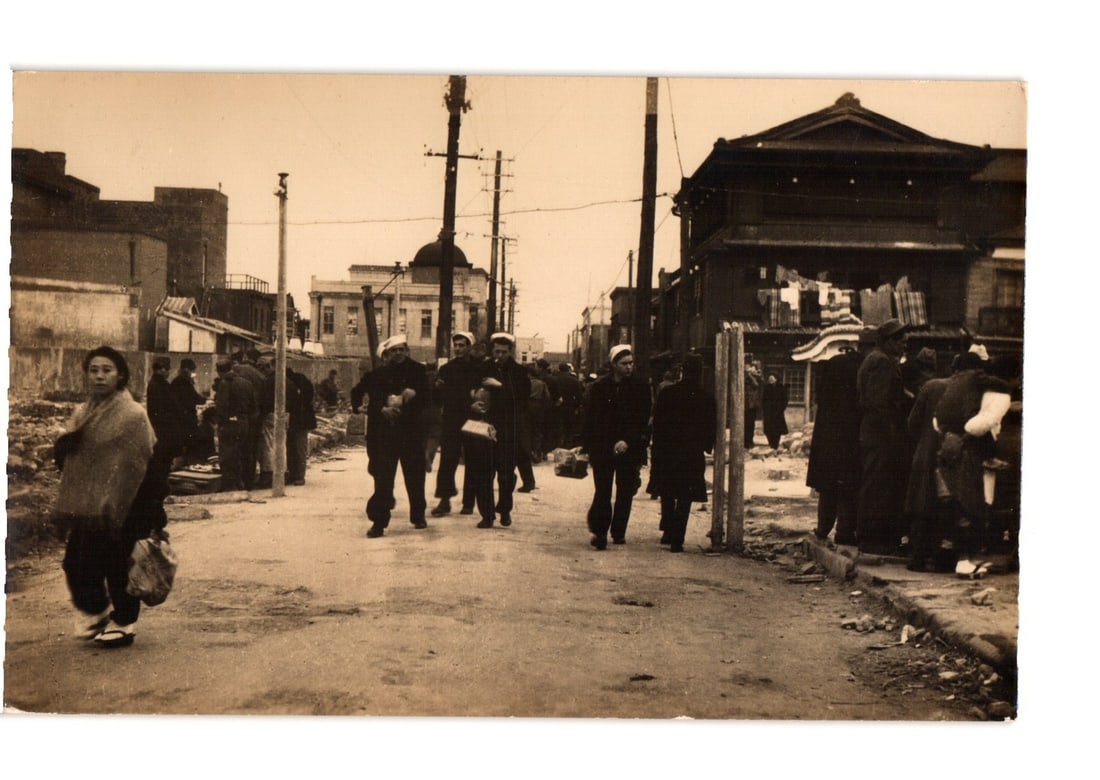 Vintage postcard: East Asian City Street Scene with Pedestrians and Uniformed Figures: A sepia-toned photographic street scene captures numerous pedestrians in an urban setting. Several individuals in the foreground and middle distance wear dark uniforms resembling sailor attire. On the