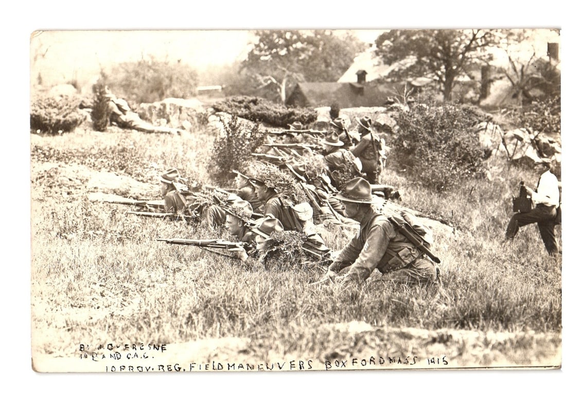 Early 20th Century Real Photo postcard showing soldiers in 1915 field maneuvers, Boxford, MA.: A sepia-toned photographic image depicts approximately eight soldiers in military uniform, positioned in a grassy field. Most soldiers are in a prone or kneeling firing position, camouflaged with brus