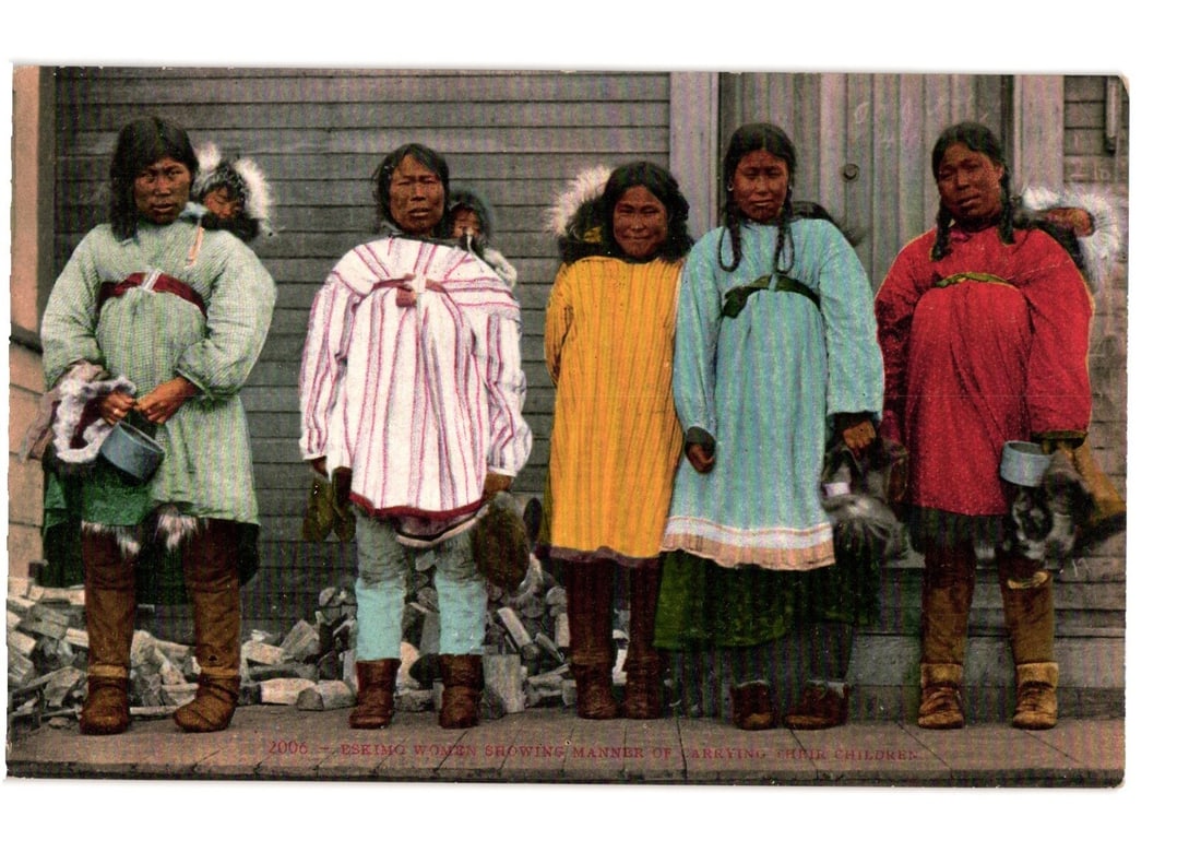 Eskimo Women Showing Manner of Carrying Children Hand-Tinted Photograph Postcard: Five Indigenous women stand facing forward in a line against a wooden wall background. The women wear varied traditional parkas, some with fur trim, in colors including light green, white and red stri