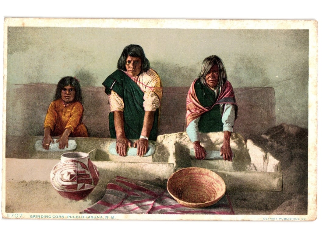 Early 20th-century postcard of Native American women grinding corn at Pueblo Laguna, N.M.: Three Native American females, two adults and one child, are depicted grinding corn. They are seated, each with their hands on a metate within a stone trough structure. The central woman wears a patte