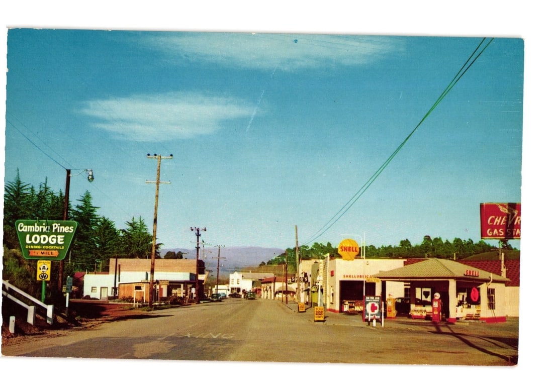 Cambria, California Street View with Shell & Chevron Gas Stations, Mid-Century Color Postcard: A street scene in Cambria, California, captured in color photography. The view looks down a main street lined with commercial buildings and utility poles under a clear blue sky with some scattered clo