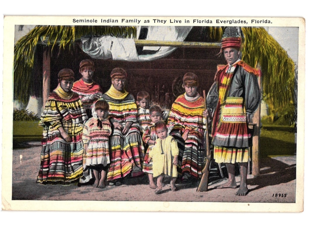 Seminole Indian Family Living in Florida Everglades, Early 20th Century postcard: A group portrait of eight Seminole individuals—three adult women, one adult man, and four children—is presented. They stand outdoors in front of a thatched-roof structure, likely a chickee. All su