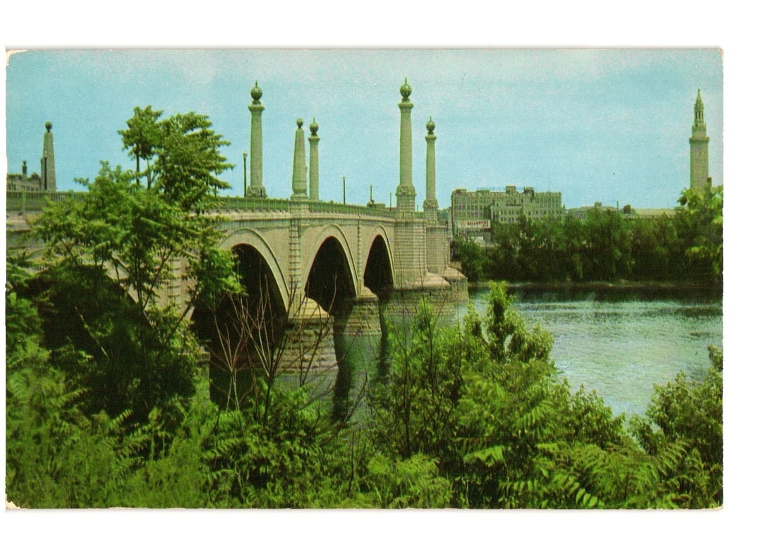 Memorial Bridge and Springfield, Massachusetts Cityscape, Mid-20th Century Color Postcard: A color photographic image features the Memorial Bridge, a stone arch structure adorned with decorative pillars, spanning a river. Lush green trees and foliage occupy the foreground, partially obscuri