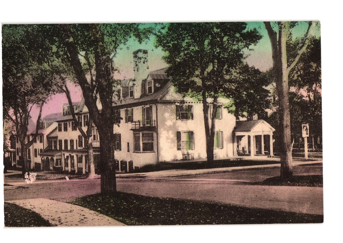Hand-colored photographic postcard of The Lord Jeffery Hotel, Amherst, Massachusetts, early 20th: A large, multi-story white building, identified as The Lord Jeffery, stands amidst numerous mature trees. The architecture features multiple dormer windows, shutters on many windows, and several porch
