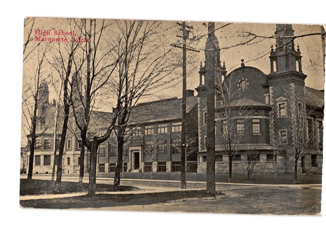 High School building in Marquette, Michigan, captured circa 1913 on a photographic postcard.: A large, multi-story institutional building, identified as the High School in Marquette, Michigan, is depicted from an oblique angle across a street. The brick structure features multiple wings, ornat