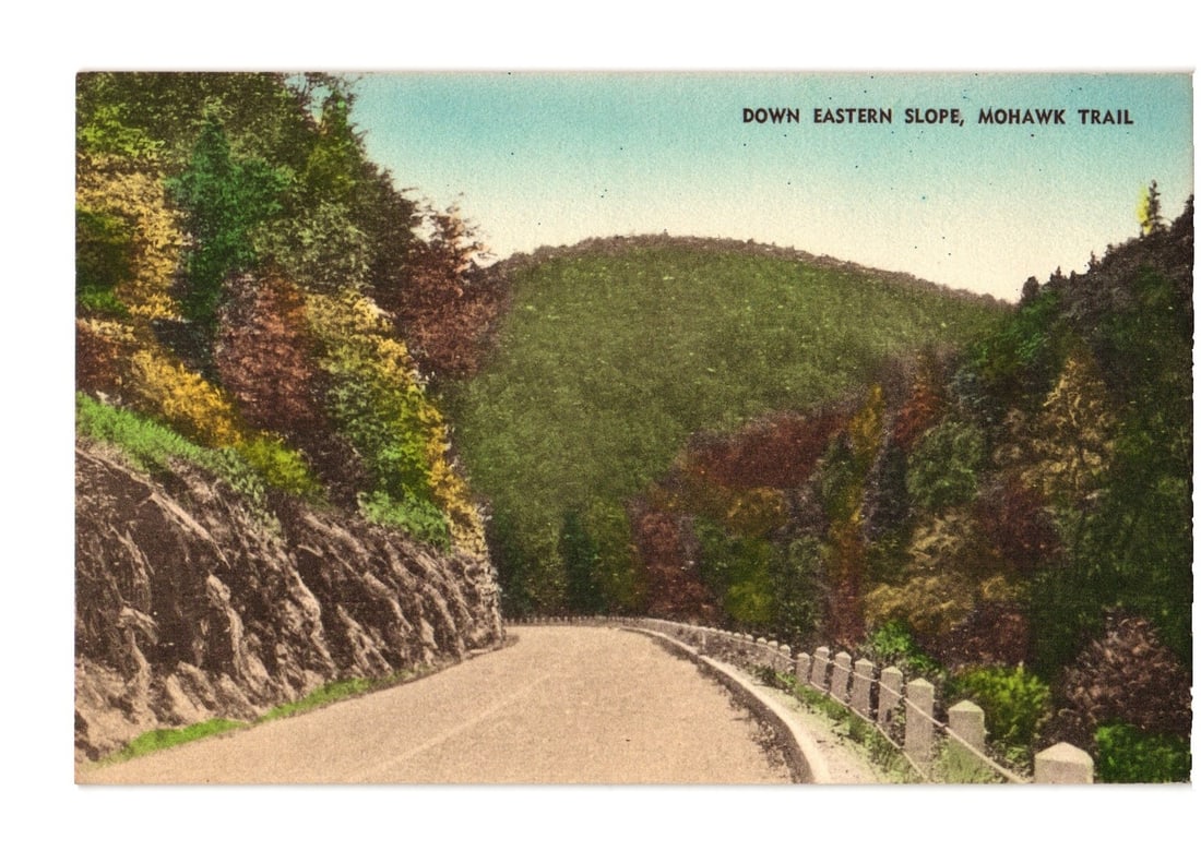 Hand-Colored postcard showing the Eastern Slope of Mohawk Trail, a scenic autumn road view.: A winding road ascends through a densely forested mountain landscape. On the left, a steep, rocky embankment borders the road, while a guardrail lines the right. The surrounding trees display autumnal