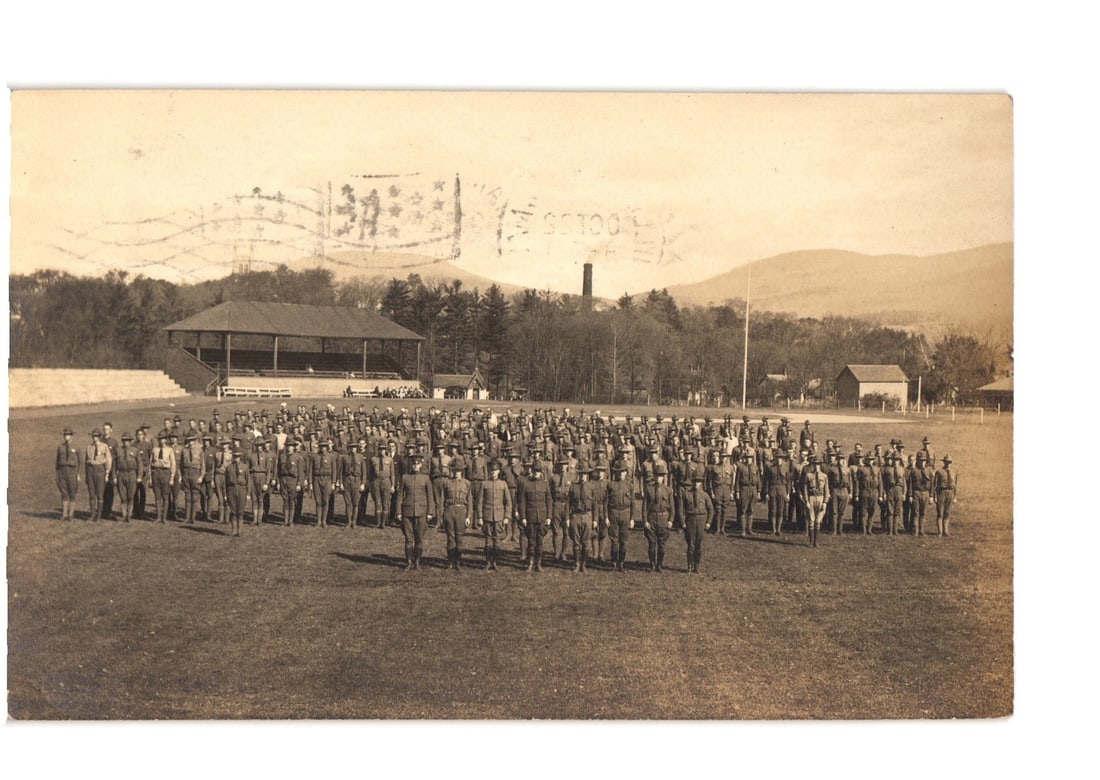 Williamstown, Massachusetts: Military Soldiers in Formation on Field, 1918 Real Photo Postcard: A large group of approximately 150 men in military uniforms stands in formation on a grassy field. The men are arranged in rows, facing forward. In the middle ground, a wooden grandstand with a roof i