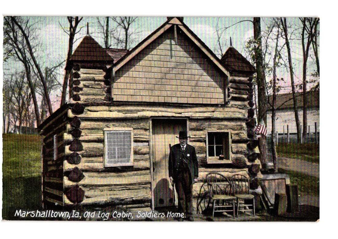 Marshalltown, Iowa: Man in Uniform at Old Log Cabin, Soldiers Home, early 20th C. postcard.: A man in a dark uniform and hat stands centrally before a rustic log cabin. The cabin, constructed of chinked logs, features a shingled gable roof with two small turret-like finials and two windows. T