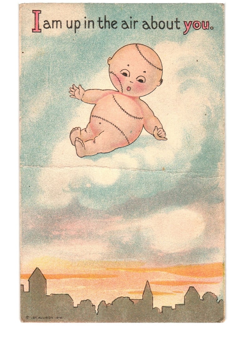 1910 Stitched Baby Floating Over Cityscape, 'I am up in the air about you' postcard: A stylized illustration depicts a bare-skinned baby or doll figure floating amidst clouds. The figure is light-skinned with stitched lines visible on its head, torso, and limbs, resembling seams. Its 