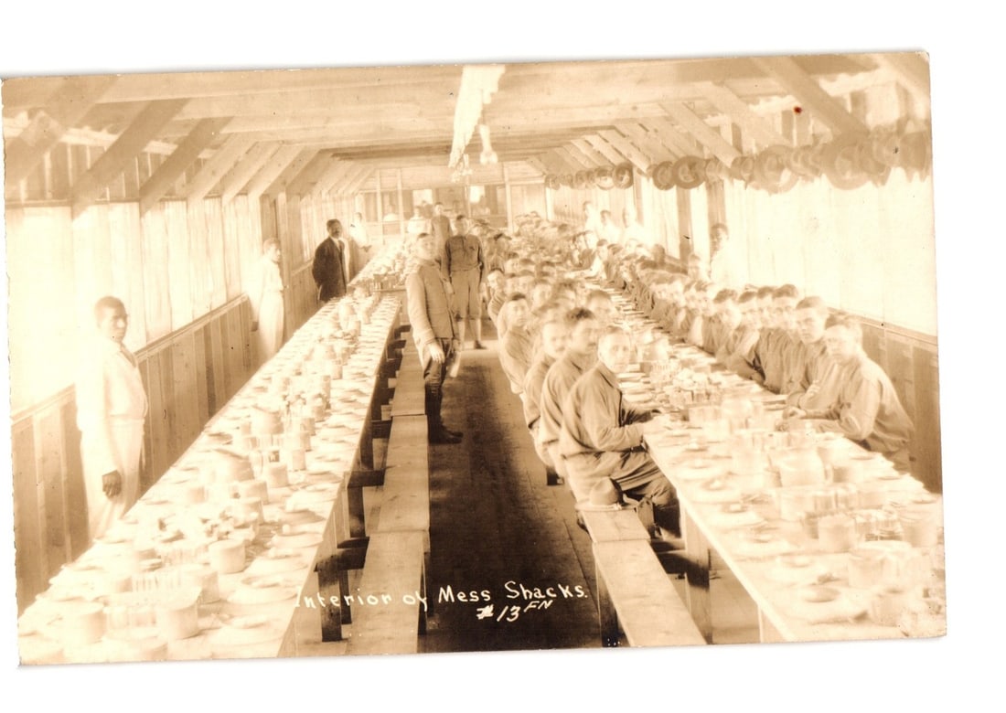 Interior View of Military Personnel Dining in Mess Shacks Postcard, Early 20th Century.: A long interior view of a mess hall or dining area. Numerous men, many appearing to be in uniform, are seated along long wooden tables, with others standing. The setting features raw wood construction
