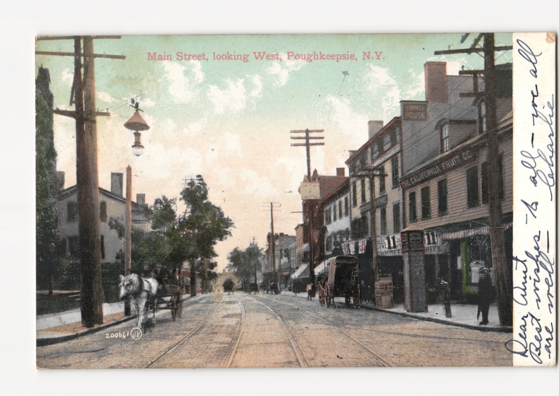 Main Street West, Poughkeepsie, N.Y. circa 1906 cityscape postcard with horse-drawn carriage: A street-level view of Main Street, looking West in Poughkeepsie, N.Y. The scene features a paved road with trolley tracks running down the center, flanked by sidewalks. Several multi-story brick and 