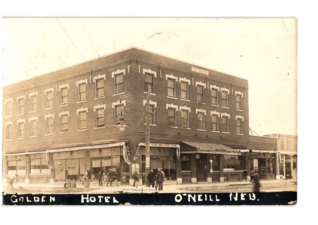 Golden Hotel, prominent brick building on street corner, O'Neill, Nebraska, circa 1920 postcard.: The Golden Hotel, a three-story brick building, stands prominently at a street corner. Commercial storefronts line the ground floor, displaying various services. Numerous windows are visible across th