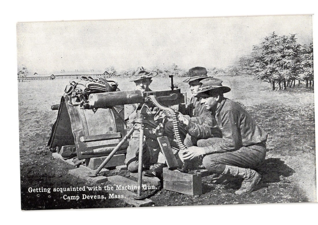 Military training postcard: Soldiers with heavy machine gun at Camp Devens, Massachusetts.: Three uniformed soldiers are depicted outdoors, operating a heavy machine gun. One soldier on the right kneels, handling the ammunition belt, while another crouches behind the weapon, seemingly adjust