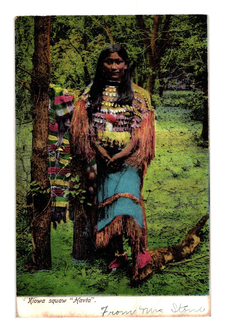 Early 20th Century Postcard: Kiowa Woman 'Navia' in Traditional Attire, Wooded Setting: A portrait-style colorized photograph depicts a young Indigenous woman, identified as Navia, standing among trees in a wooded landscape. She faces forward, looking directly at the viewer. She wears a 