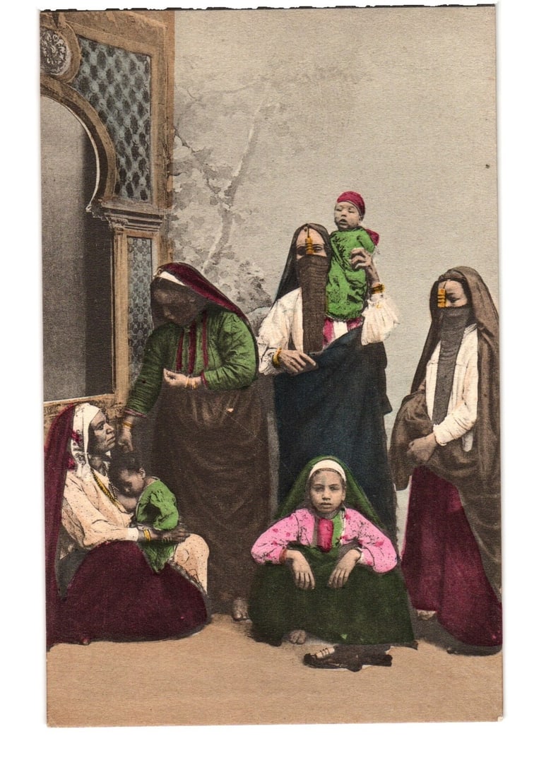 Early 1900s Egypt: Group of Arabian Women and Children in Traditional Attire Postcard: A group portrait depicts five women and two children in traditional attire within an interior setting. Two women stand on the right, one holding an infant wrapped in green cloth. Another woman stands 