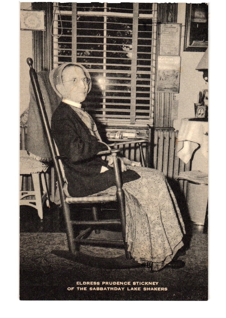 Eldress Prudence Stickney of Sabbathday Lake Shakers, seated portrait postcard, mid-20th century.: A black and white photographic postcard features Eldress Prudence Stickney of the Sabbathday Lake Shakers. She is seated in a wooden rocking chair, positioned against a backdrop of a room with Venetia