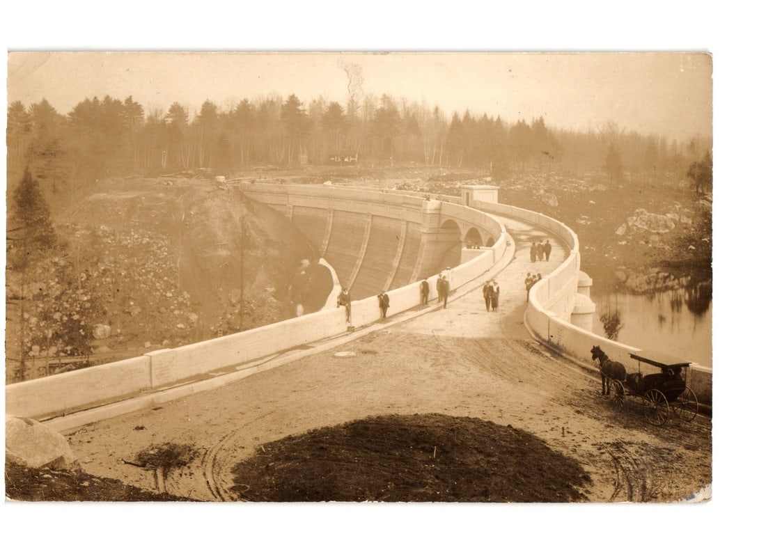 Sepia Photo Postcard: Concrete Dam Construction, Horse-Drawn Cart, Forested Landscape, Early 20th: A large, curved concrete dam is depicted, appearing to be under construction or recently completed. The dam features a wide walkway or road on its upper surface, where approximately eight individuals 