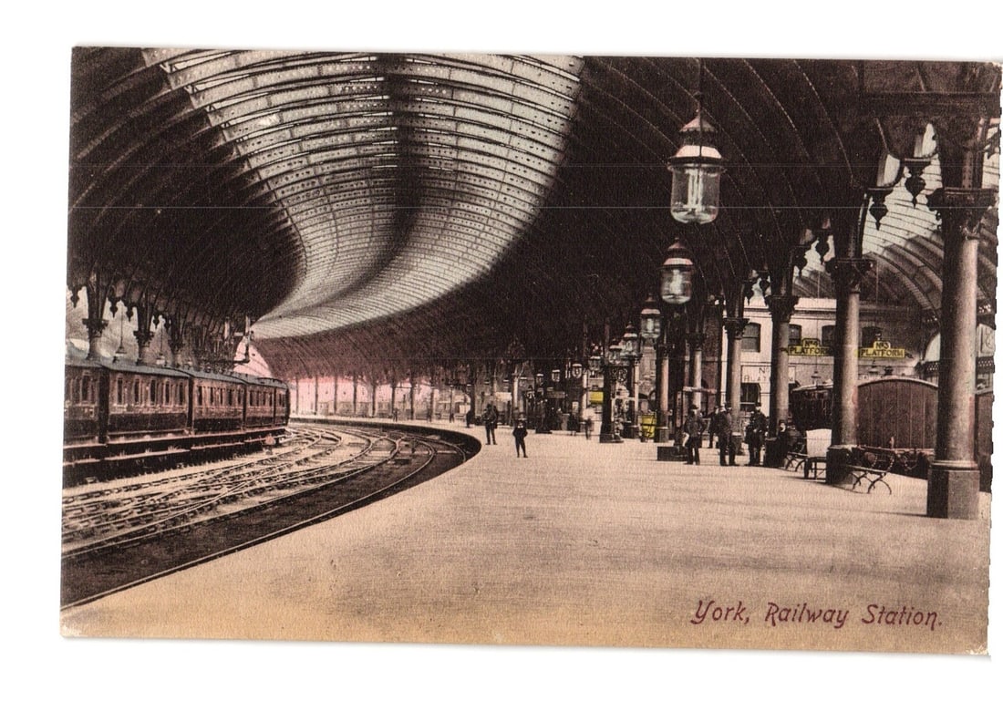 York Railway Station Interior View with Train and Travelers, Early 20th Century postcard.: The interior of a large railway station is depicted, featuring an arched glass and steel roof spanning over multiple tracks and a wide platform. A train with several carriages is visible on the tracks