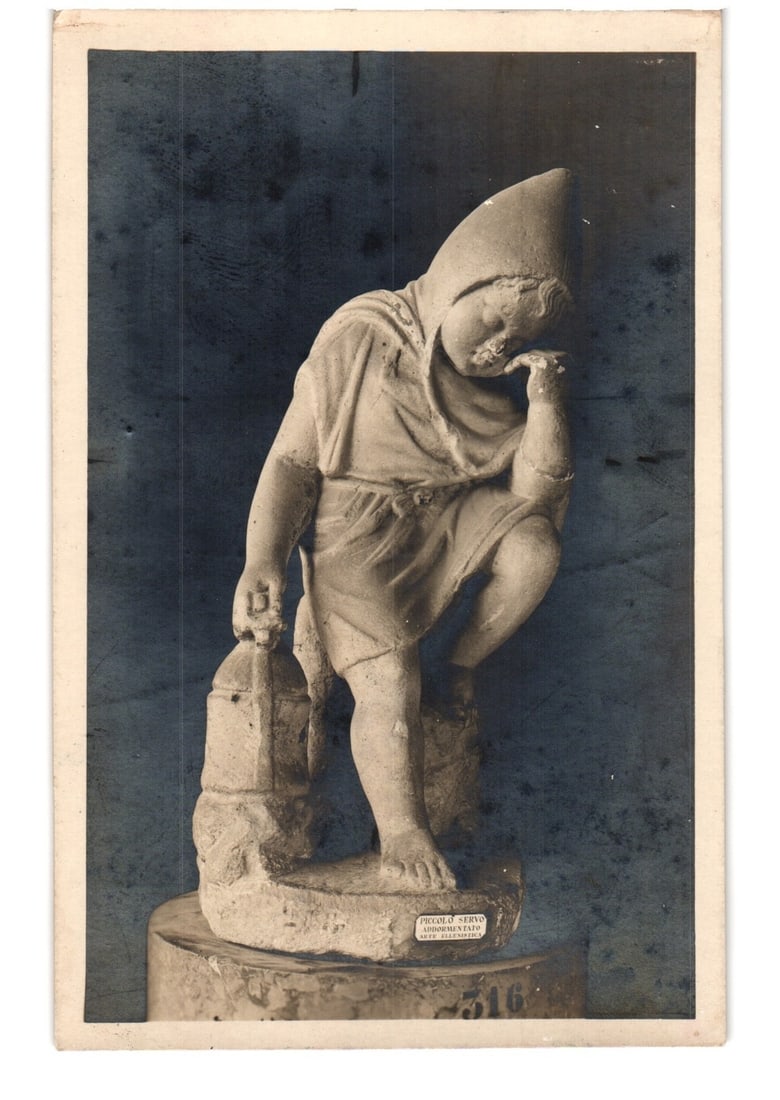 Photographic postcard of the Hellenistic statue 'Sleeping Little Slave' from Roman National Museum.: A full-length photographic image depicts a statue of a young boy, possibly a putto or eros, with a pensive expression, wearing a conical hat and draped garment. He is seated, leaning his head on his r