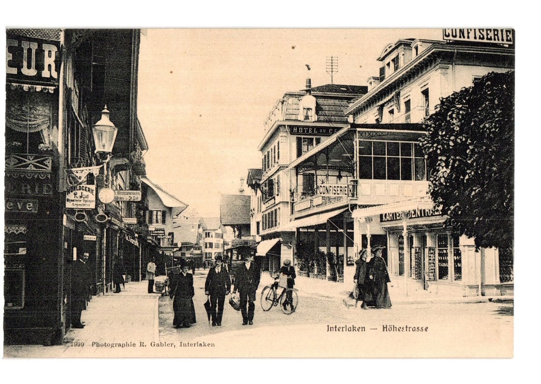 Interlaken, Switzerland Hohestrasse Street View with Pedestrians and Shops Photographic Postcard: A street scene depicts Hohestrasse in Interlaken, Switzerland, with several pedestrians and commercial buildings. Men in suits and hats and women in long dresses and hats walk on the street; one perso