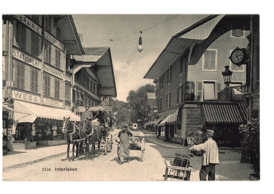 Early 20th-century street scene postcard with shops, pedestrians, and horse carriages, Interlaken.: A sepia-toned photographic street scene depicting a bustling town square or main thoroughfare. On the left, multi-story buildings house shops with signs reading "LIBRAIRIE", "LUES SUISSES", "STATIONER