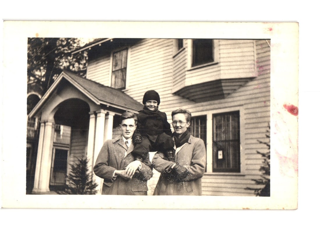 Black and white photographic postcard depicting a family portrait in front of a house, RPPC.: A man, a woman, and a young child are positioned outdoors in front of a large residential house. The child, wearing a dark winter hat and coat, sits on the man's shoulders. The man, in a collared shir