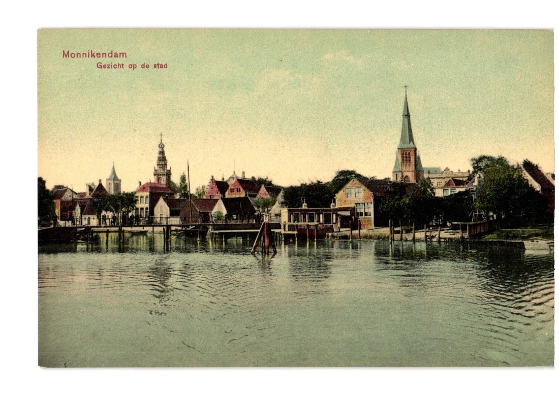 Monnikendam, Netherlands: City view across water with prominent church spires, 1908 chromolithograph: A picturesque panorama of Monnikendam, Netherlands, captured across a tranquil body of water. The town's skyline features numerous buildings with gabled roofs, punctuated by two distinct towers. On th