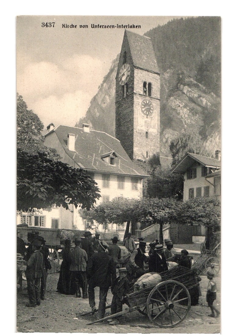 Early 20th Century postcard: Unterseen-Interlaken Church with an active market scene.: A prominent church tower featuring multiple clock faces rises above surrounding buildings and an active outdoor market scene. The tower, with a dark, pointed roof, is centrally positioned against a ba