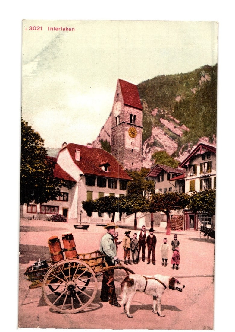 Early 20th Century Interlaken street scene postcard featuring man, dog, cart, village, clock tower.: A colorized photograph depicts a village street scene in Interlaken. In the foreground, a man wearing a hat stands next to a two-wheeled wooden cart laden with barrels. A dog on a harness stands besid