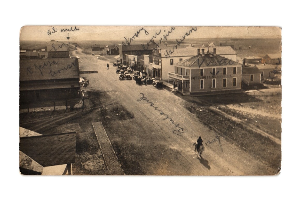 Street scene with Hotel Thorburn, automobiles, and horseback rider, Thorburn, Saskatchewan postcard.: An elevated photographic view depicts a main street in a rural town. In the foreground, a person on horseback rides away from the viewer on a dirt road. Several early model automobiles are parked alon