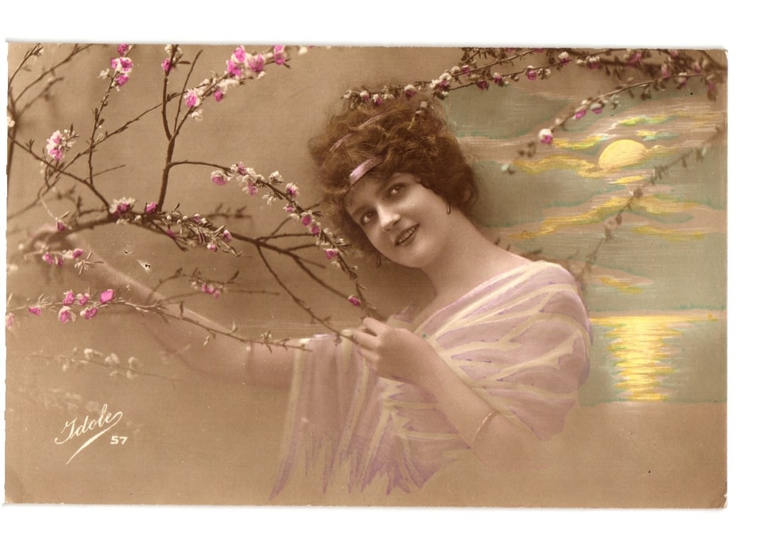 Early 20th Century 'Idole' Postcard of Woman with Blossoms and Stylized Sunset: A young woman with dark, curly hair and a subtle smile poses next to flowering branches adorned with pink blossoms. She wears a light, draped garment, possibly a shawl, in a pale lavender hue. Her gaz
