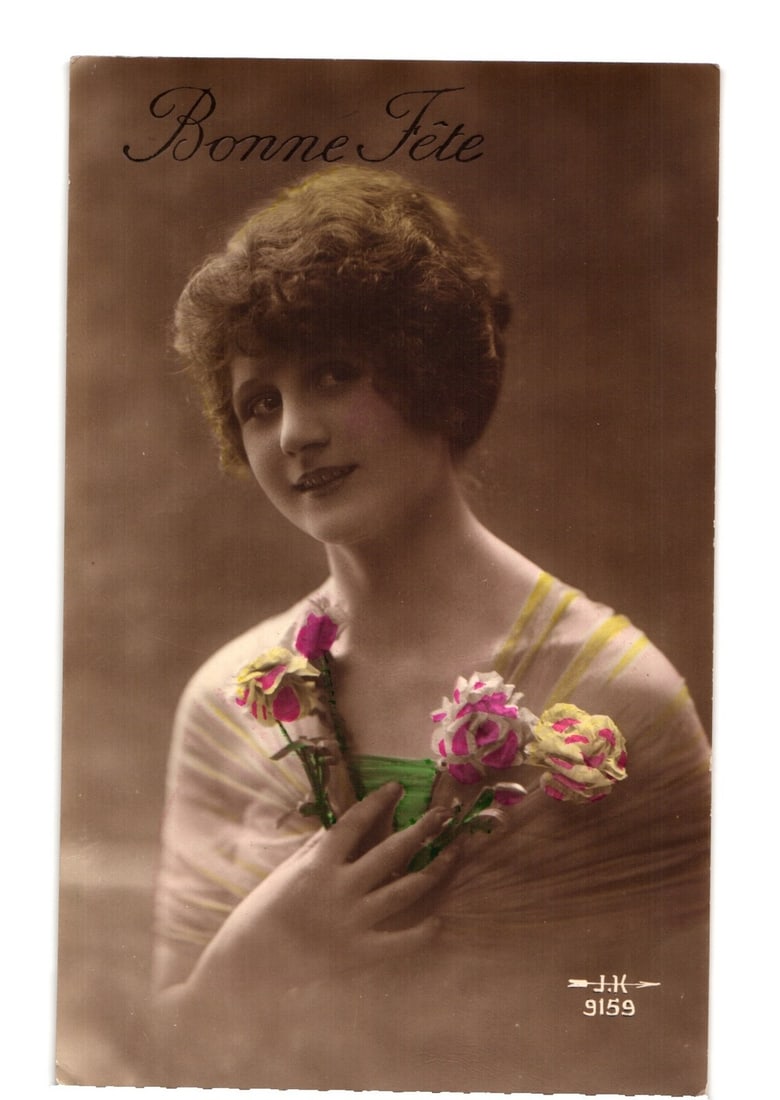 Young Woman Holding Flowers 'Bonne Fete' Hand-Colored Photographic Portrait Postcard, 1910s: A young woman is depicted in a sepia-toned, hand-colored photographic postcard, presented in a portrait orientation. She is shown from the waist up, facing slightly left but looking directly at the vi