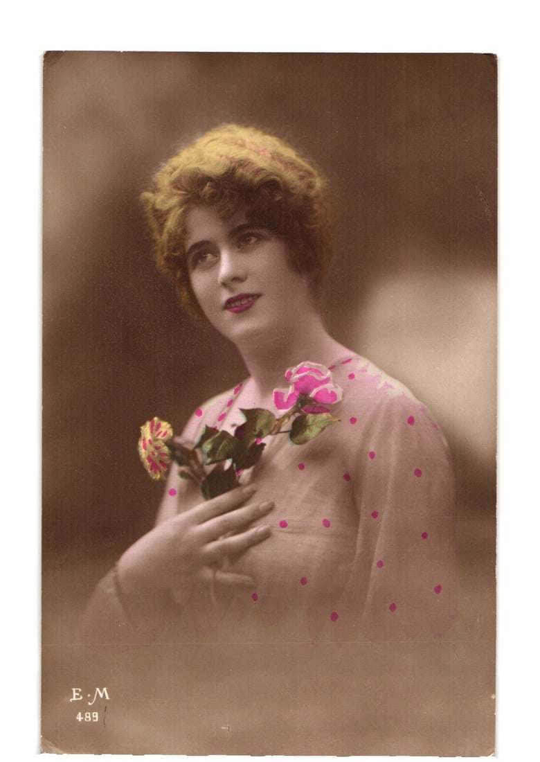 Hand-colored portrait postcard of a young woman with pink flowers, French Made, early 20th century.: A hand-colored photographic portrait features a young woman posed waist-up, facing the viewer's right and looking upwards. She has short, wavy, light-colored hair and red lips. She wears a sheer, ligh