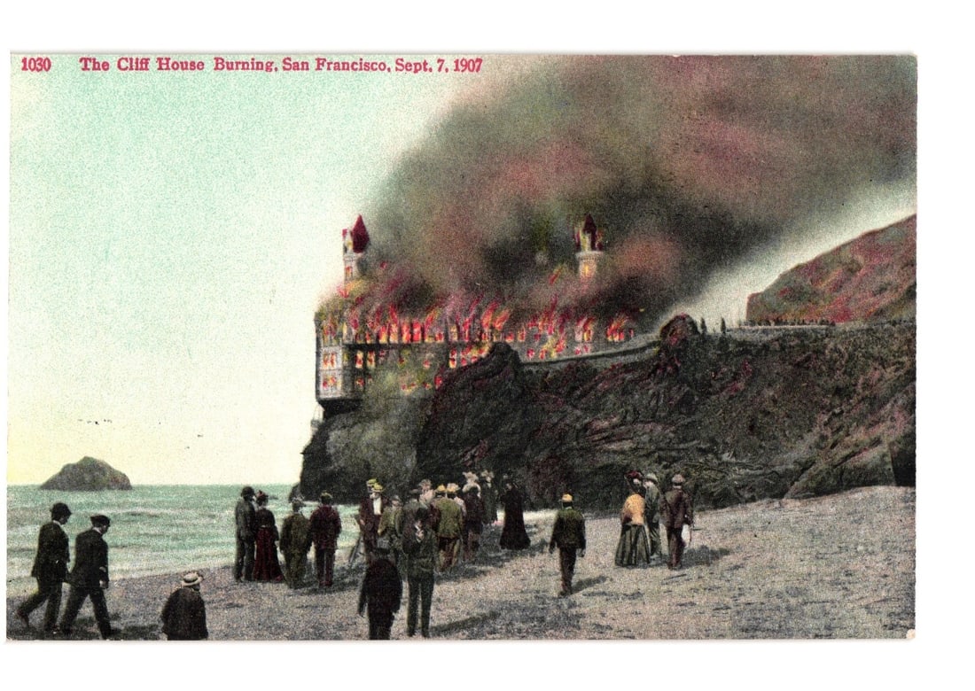 Panoramic lithograph postcard depicting the Cliff House burning in San Francisco, September 7, 1907: A multi-story building, identified as The Cliff House, is depicted in flames, with heavy dark smoke rising into the sky. The building is situated on a rocky cliff overlooking the ocean. Numerous indiv