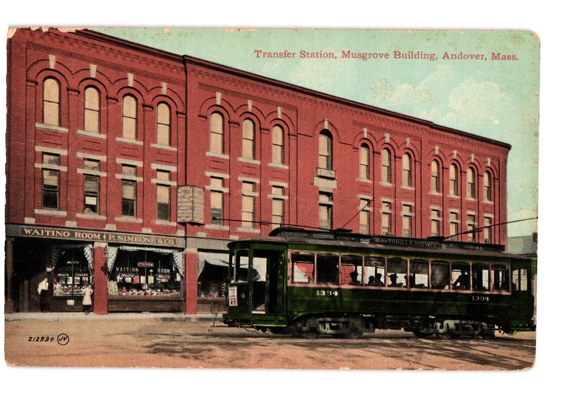 Colorized postcard of Transfer Station, Musgrove Building, Andover, Massachusetts, featuring a: A street scene depicts a large, three-story red brick building identified as the Musgrove Building, featuring arched windows on its upper floors. The ground floor includes a storefront with signage re