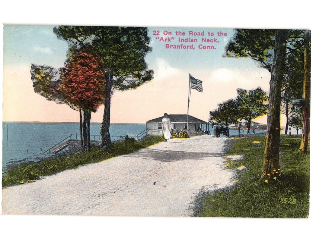 Scenic postcard of the Road to 'Ark' Indian Neck, Branford, Conn., early 20th century.: A winding road leads towards a building situated at the edge of a body of water. A woman in a white dress walks on the road in the foreground. Trees with green and reddish-orange foliage frame the pat