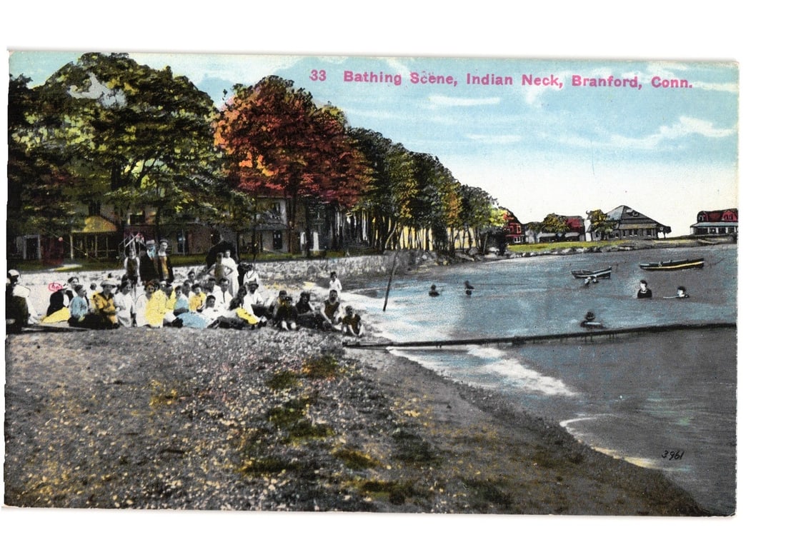Bathing Scene at Indian Neck, Branford, Connecticut, early 20th-century colorized postcard: A bustling bathing scene depicts numerous individuals on a pebbled and sandy shore. Several bathers are wading or swimming in the water, while others are seated or standing on the beach. Large trees w