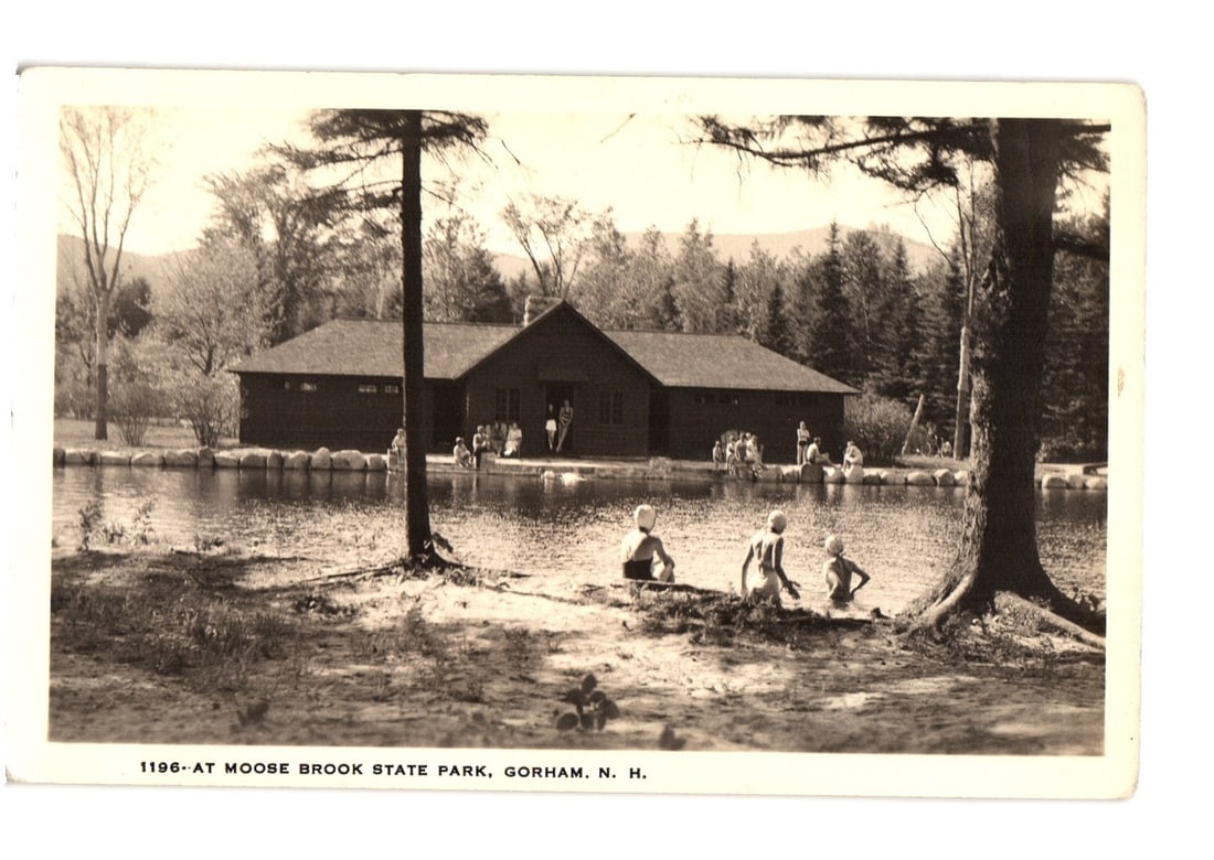 Vintage Postcard of Moose Brook State Park, Gorham, N.H., featuring a building and people.: A scenic view of Moose Brook State Park, depicting a large park building situated along the bank of a pond or lake. Numerous individuals are visible; several adults are gathered near the building's en