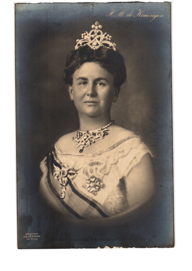 H. M. the Queen, formal photographic portrait postcard by F. A. Dénn, The Hague.: A formal photographic portrait depicts a woman adorned with a tiara, pearl necklace, and an ornate sash bearing medals, presented in a bust-length composition against a dark background. The image is a
