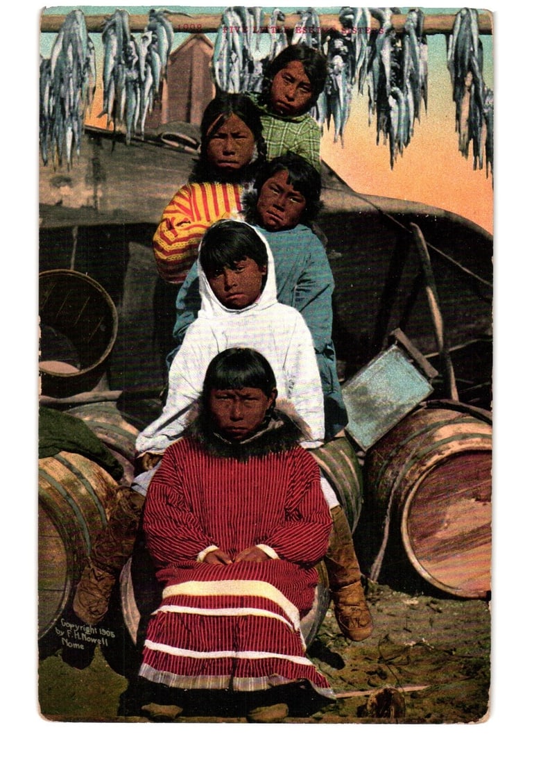 Hand-colored Postcard: Five Indigenous Children with Drying Fish, Barrels, circa 1908: Five Indigenous children are depicted in a hand-colored photographic image, arranged vertically in a group. The foreground child is seated, wearing a striped red and white tunic with a dark collar. Be