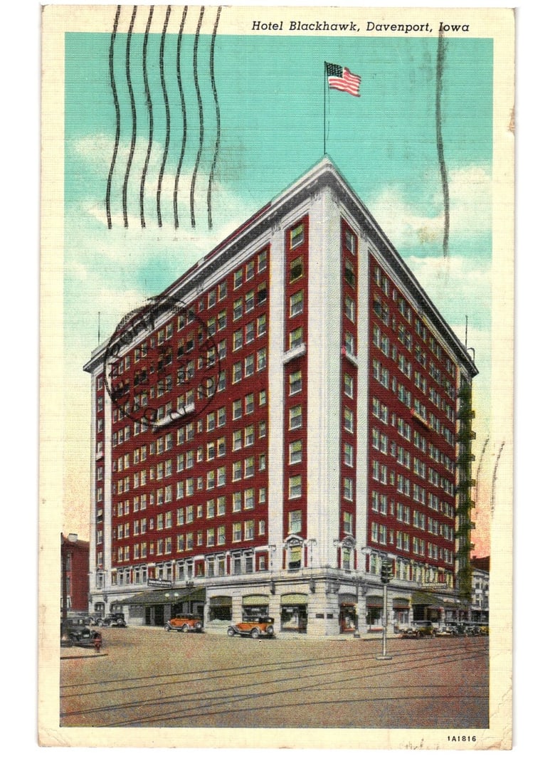 1941 Davenport, Iowa City Scene Postcard: Hotel Blackhawk, a large corner building.: A street-level view of the Hotel Blackhawk, a multi-story building with a red brick facade and white vertical architectural elements, located in Davenport, Iowa. The hotel stands prominently at a stre